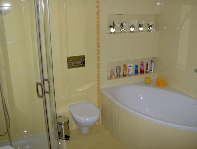 Jasna łazienka z narożną wanną, kabiną prysznicową, toaletą i półkami w ścianie z dekoracjami i kosmetykami, wykończona żółtymi płytkami.